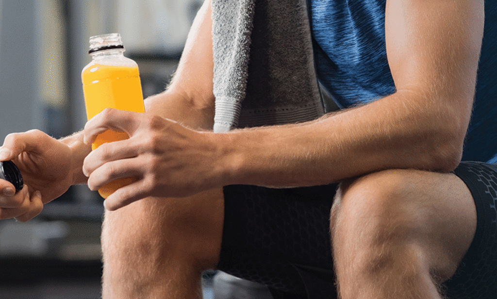 Man sitting in a gym, in workout clothes, towel draped on his shoulders and holding an energy drink.