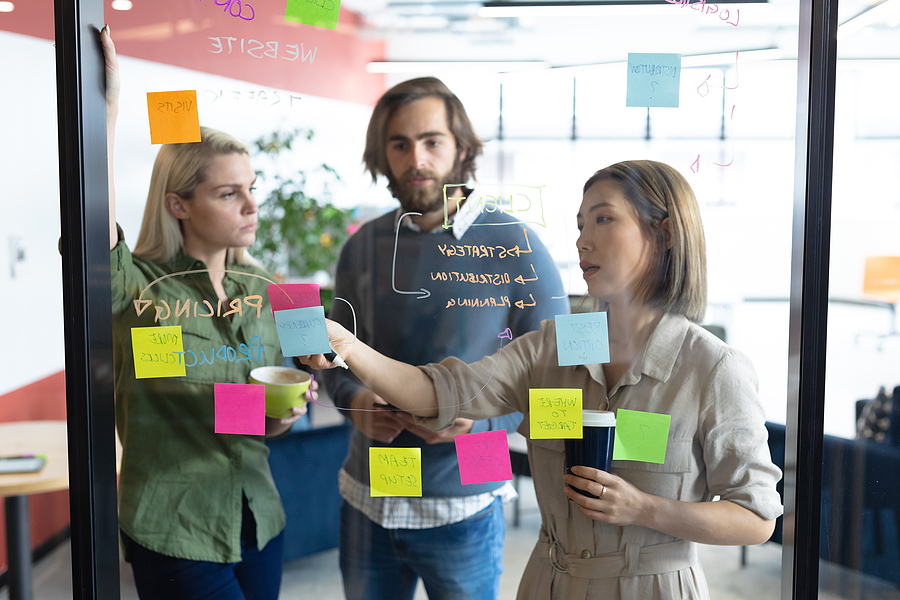 Three people standing by a glass wall adding sticky notes to it and discussing business plans